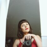 Woman in red top takes a selfie with a DSLR camera, reflecting in mirror.